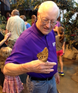 A man in a purple shirt is looking at a butterfly that has landed on his hand while other people are in the background.