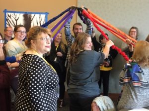 A group of attendees are gathered in a circle holding colorful bands that a man in the middle is holding one end of.