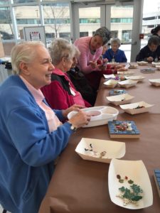 A group of women are seated at a table with trays of art supplies. The women in the front is smiling and choosing objects from a tray.