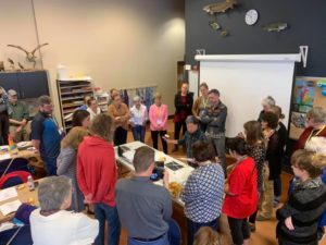 An artist is standing at a table over supplies while a group of people are standing around table watching and listening.